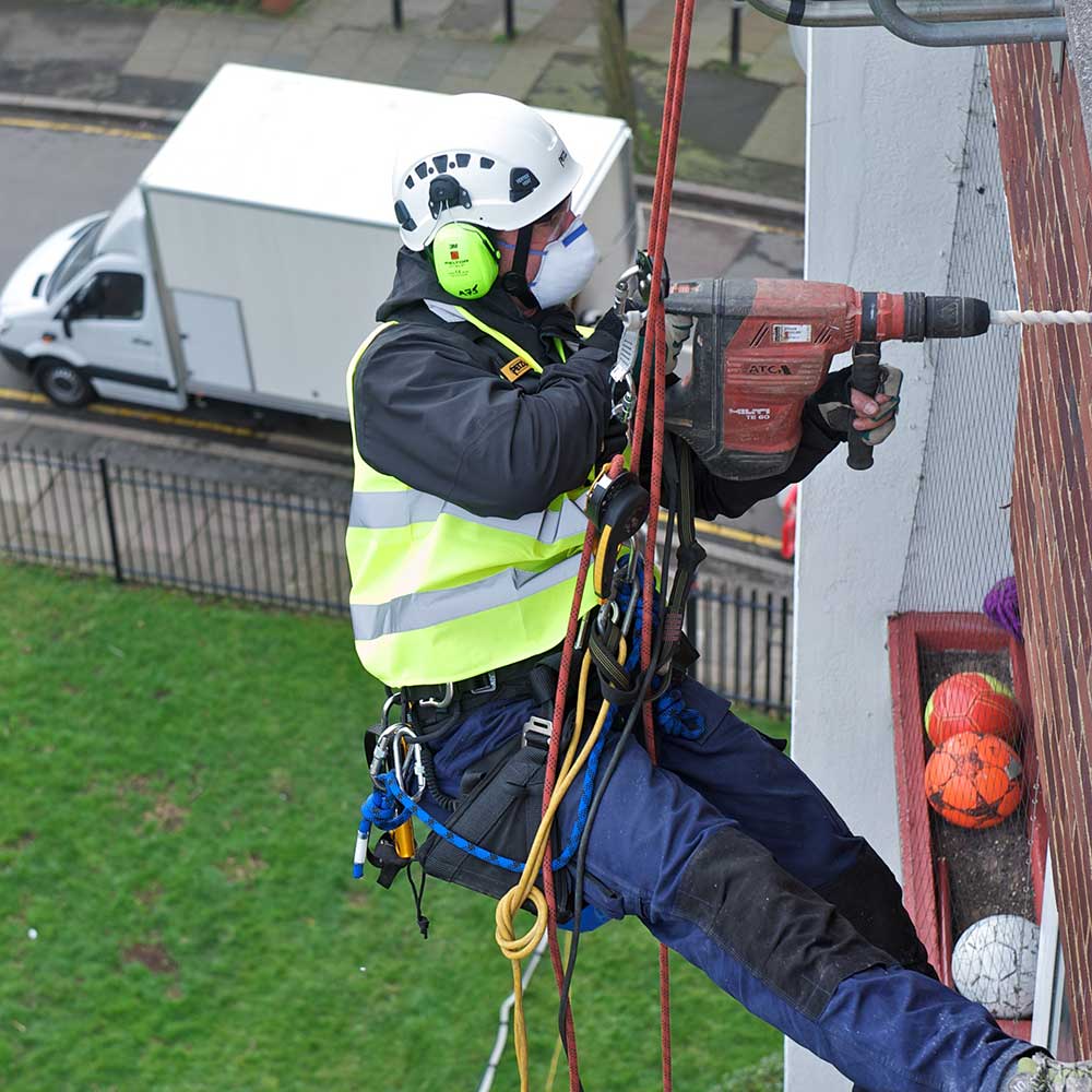 Axis person using a large drill whilst abseiling down the side of a building