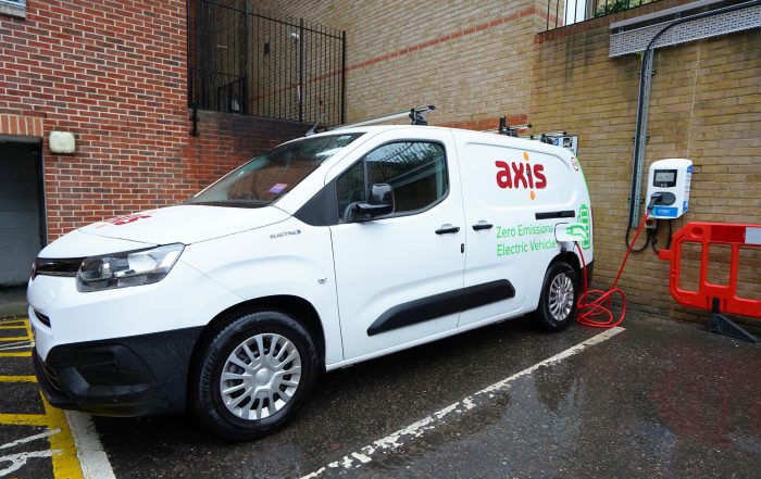 White van with black windows and wheels. Red text with Axis logo. Van on a parking street. Small square of electric charging point behind the vehicle on a light brown brick wall.