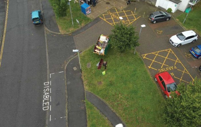 Bird's eye view of grey roads, green grass an a skip. Block of flats on the right.