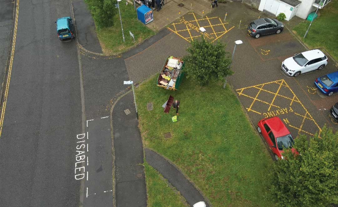 Bird's eye view of grey roads, green grass an a skip. Block of flats on the right.