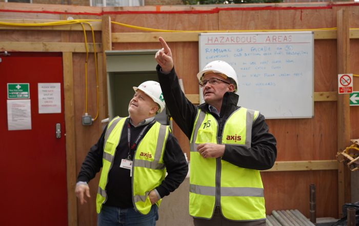 Two men in yellow high viz jackets and hard hats on a site with wooden hoarding