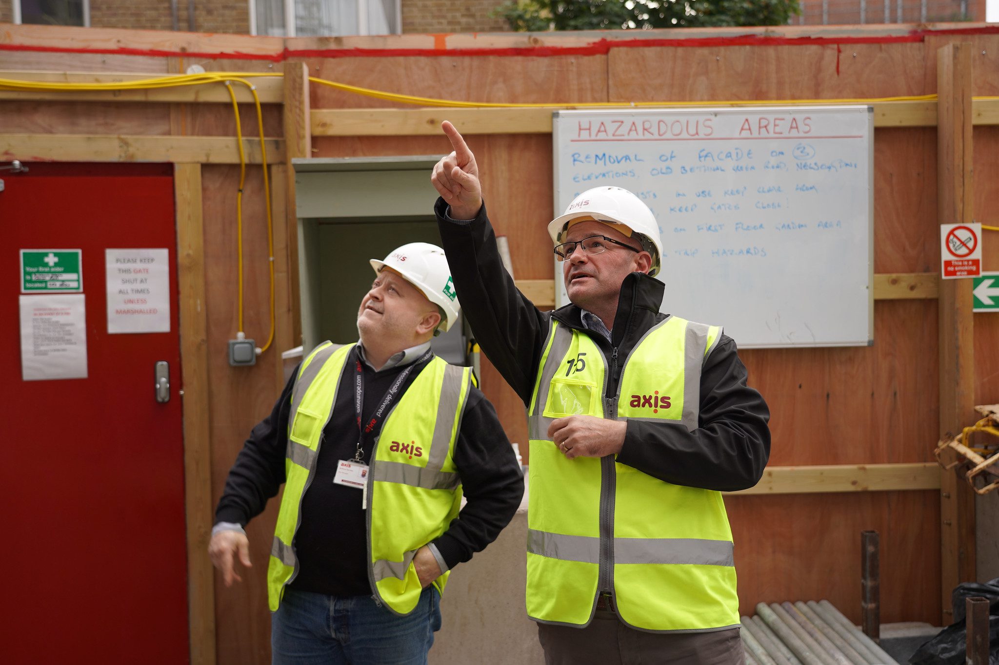 Two men in yellow high viz jackets and hard hats on a site with wooden hoarding