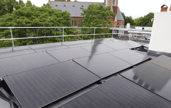Black solar panels on a black roof with a white chimney. Building and trees behind