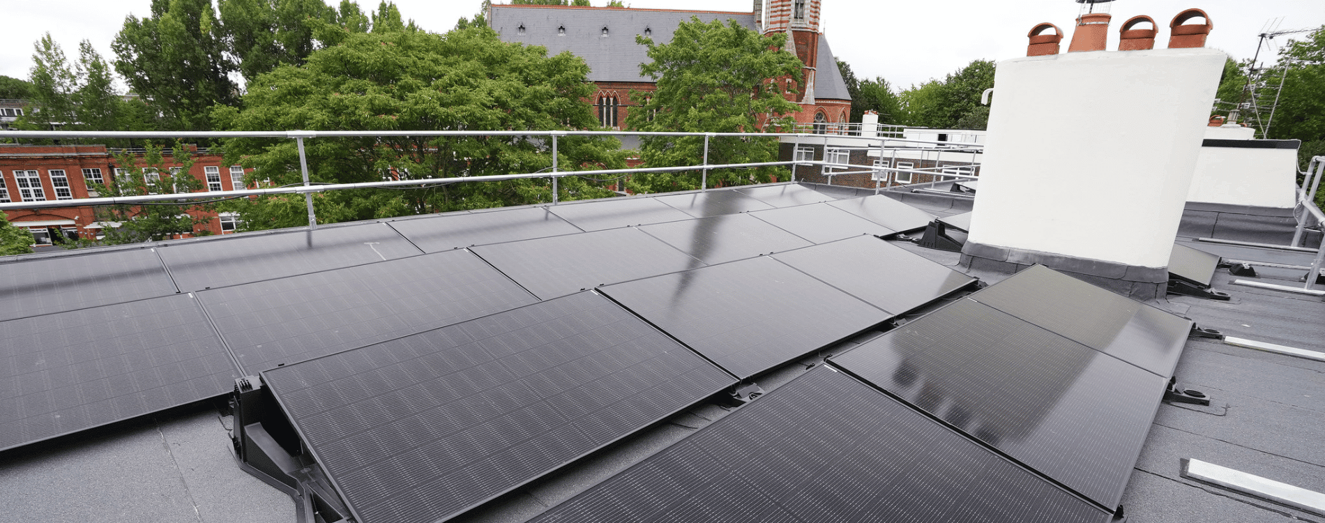 Black solar panels on a black roof with a white chimney. Building and trees behind
