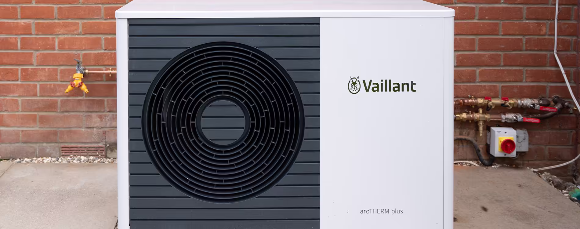 A Vaillant aroTHERM plus air source heat pump outdoor unit, professionally installed against a red brick wall of a house.