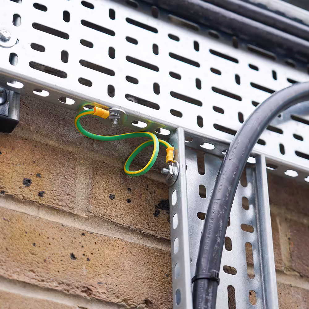 A close-up of a green and yellow earth bonding wire correctly installed on a galvanised steel cable tray, highlighting a crucial electrical safety detail.