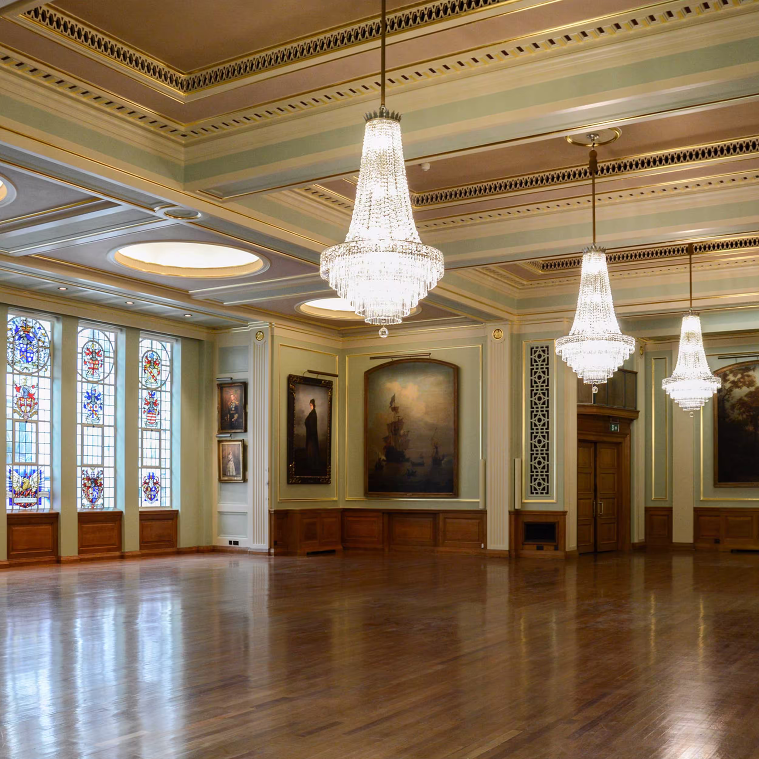 A wide view of a magnificently restored historic hall, emphasizing the vast polished wood floor, hanging crystal chandeliers, and large paintings in an elegant event space.