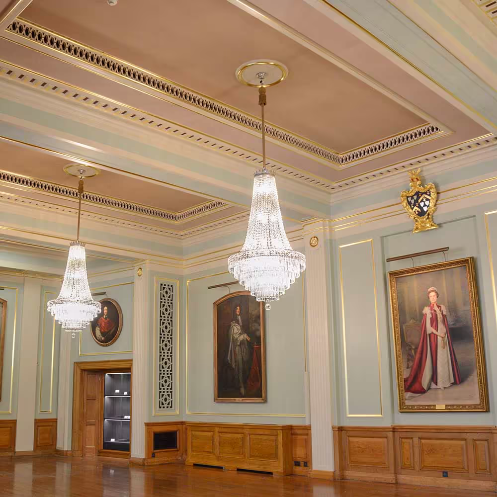The interior of a restored grand hall, featuring large crystal chandeliers hanging from a high, ornately decorated ceiling above historic royal portraits on the walls.