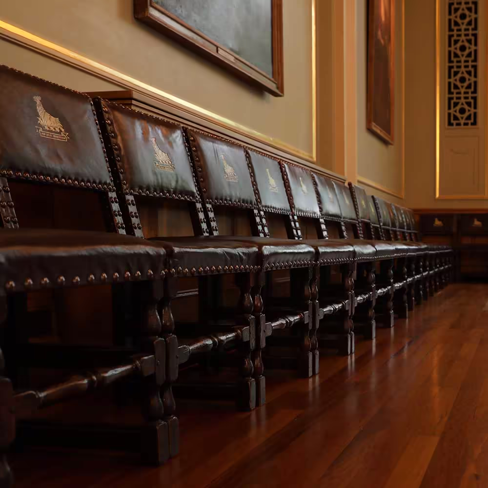 A row of restored antique dark wood and leather chairs with an embossed crest, showing the quality of specialist furniture restoration within a historic hall.