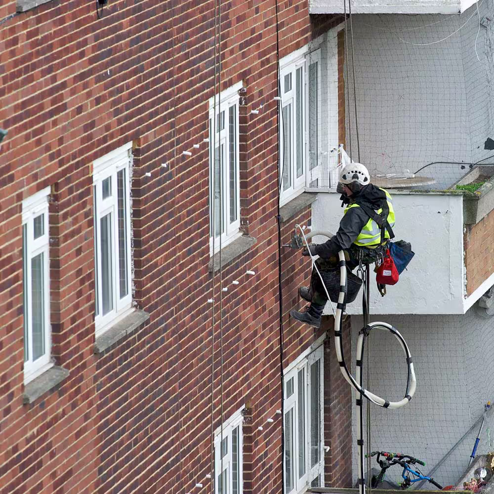A close-up of a technician abseiling down a brick wall while injecting insulation material through a hose into pre-drilled holes.