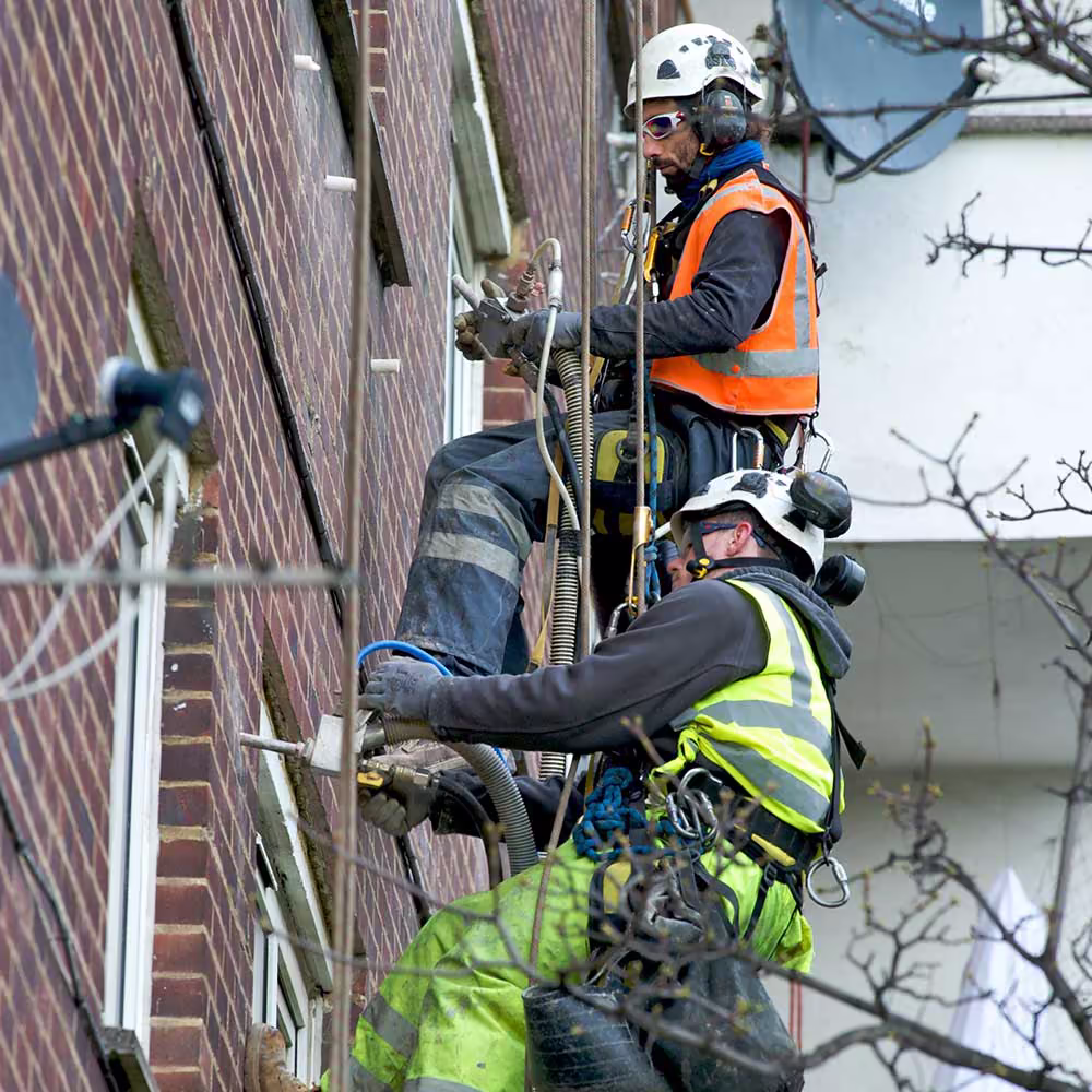 Two rope access technicians work together as a team while suspended on a high-rise building, drilling holes for cavity wall insulation.