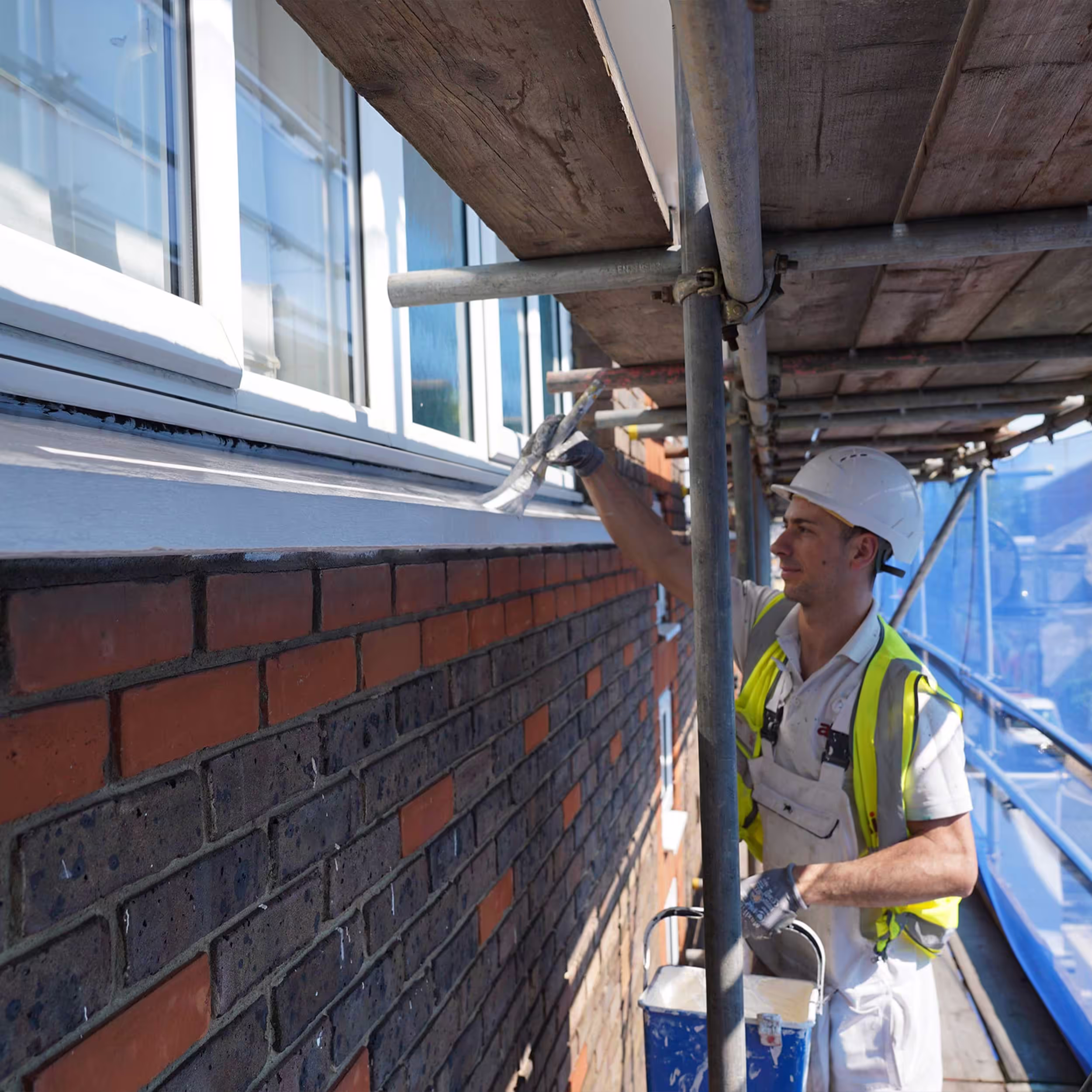 A professional painter in a hard hat and safety vest works from scaffolding, carefully painting the exterior woodwork of a brick building during communal repairs.