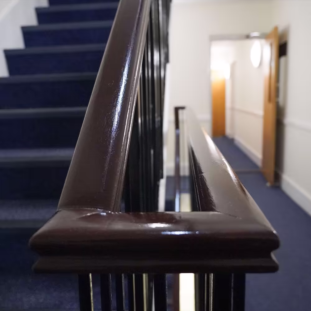 A close-up of a freshly varnished dark wood banister in a communal stairway, showing the high-gloss finish next to newly laid blue carpet.