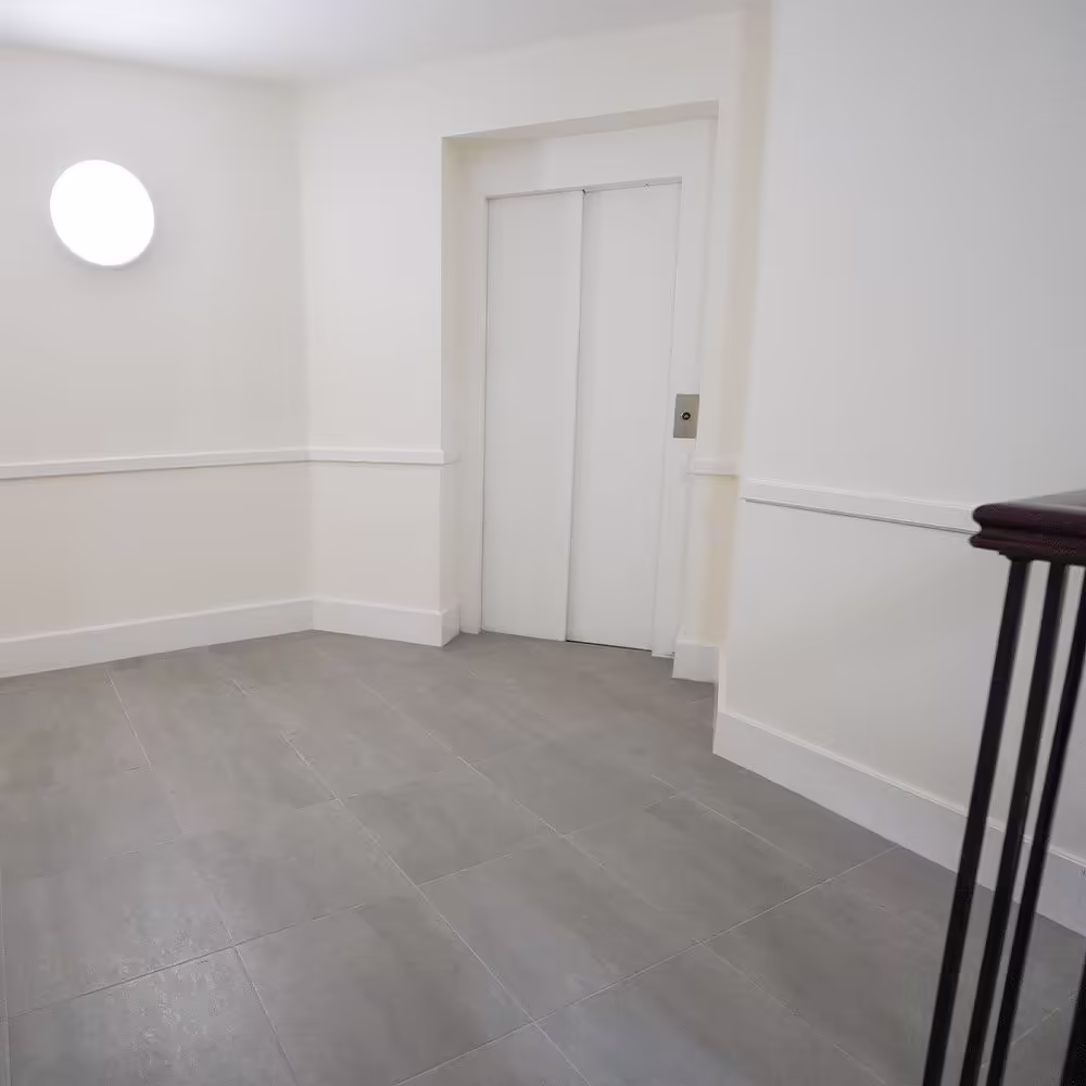 A redecorated communal landing with pristine white walls, new grey tiled flooring, a modern circular wall light, and a lift entrance.