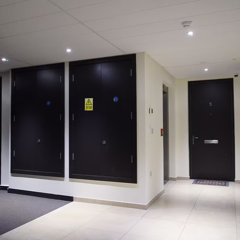 A smart, newly decorated communal hallway featuring crisp white walls, light-coloured floor tiles, and contrasting dark-painted flat entrance and service cupboard doors.