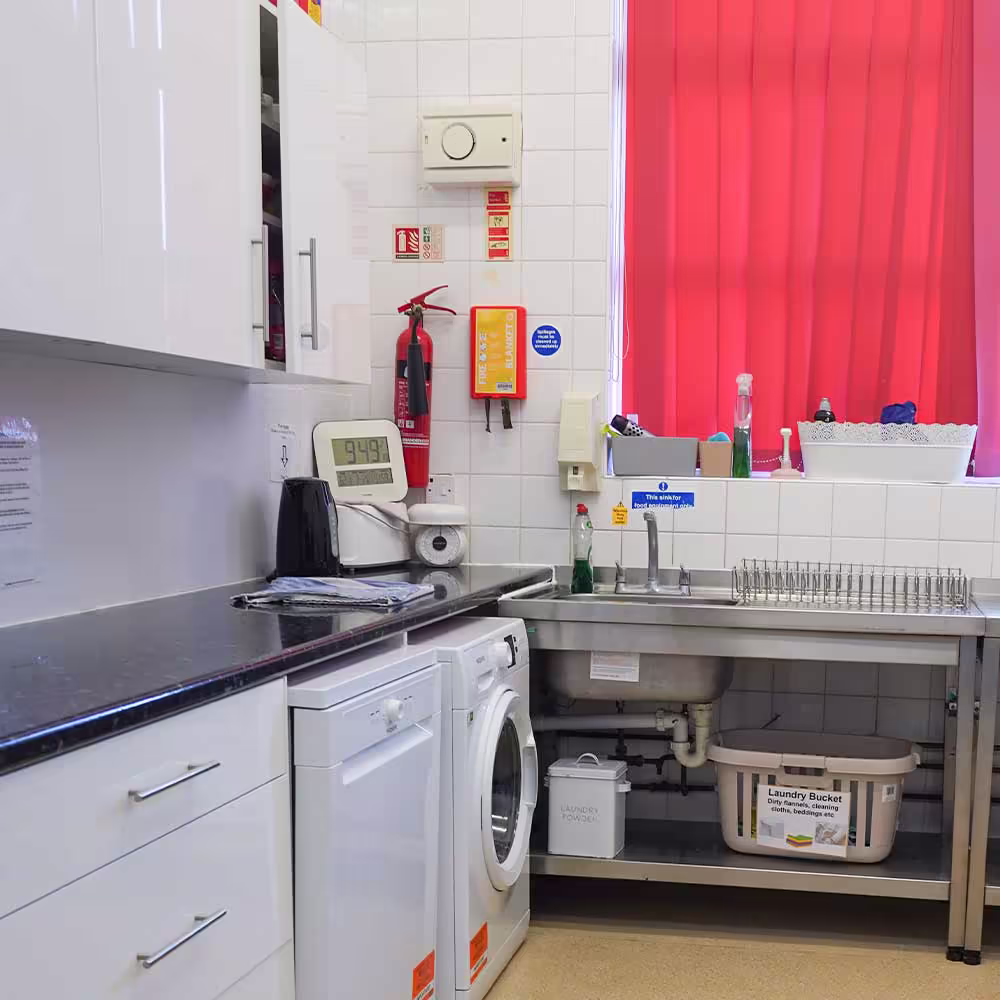 The clean and functional redecorated kitchen and utility area in a community centre, featuring new cabinets, a washing machine, and essential safety equipment.