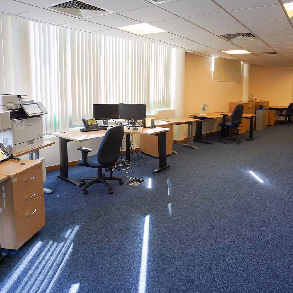 The refurbished community centre office with new blue carpet, modern desks equipped with computers, and bright light coming through the window blinds.