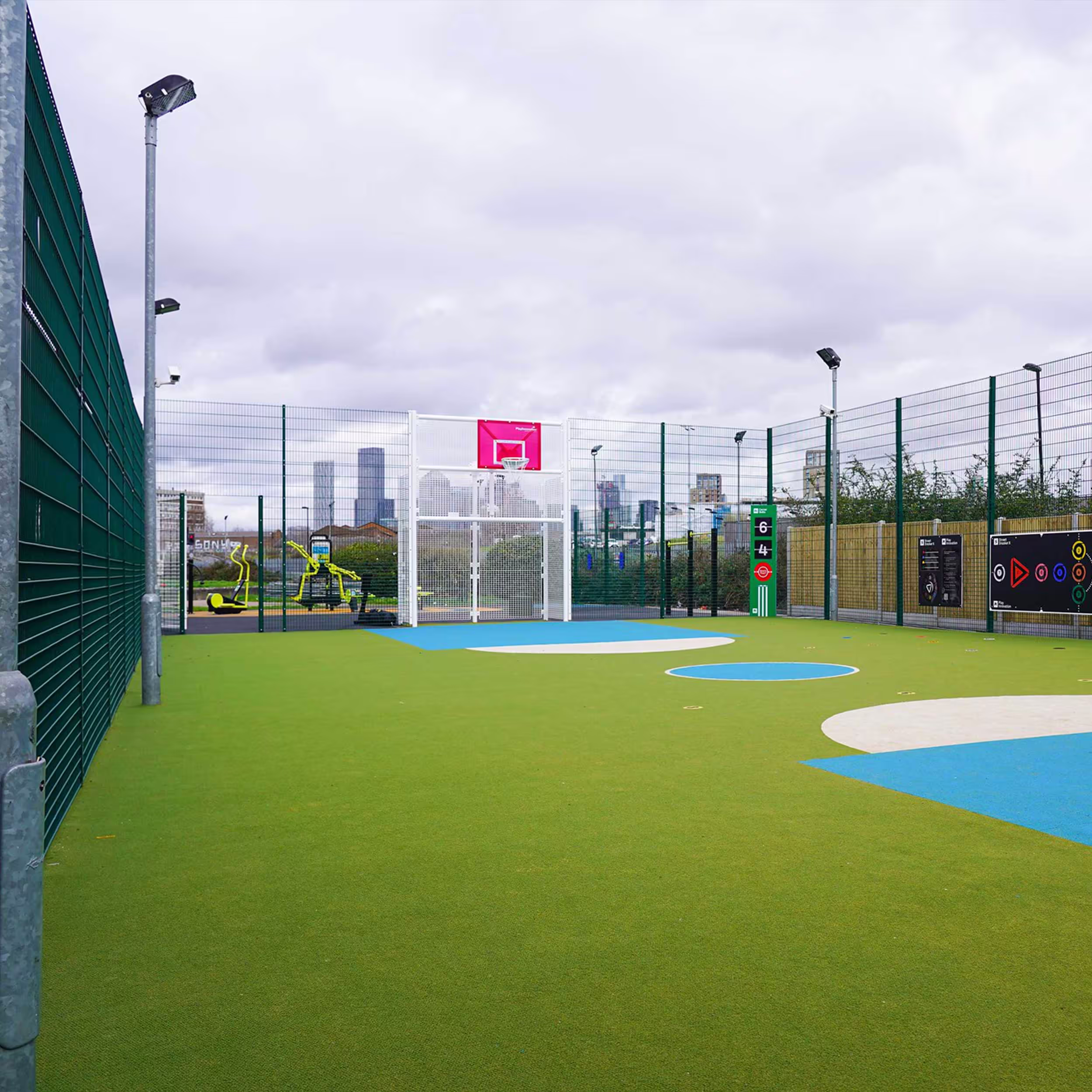 A modern, urban multi-use games area (MUGA) with an artificial grass pitch, basketball hoop, and goal, set against a city skyline.