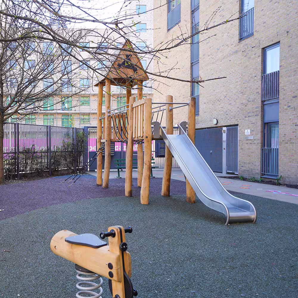A natural wood adventure playground next to a modern apartment block, featuring a timber climbing frame with a slide and a spring rocker.