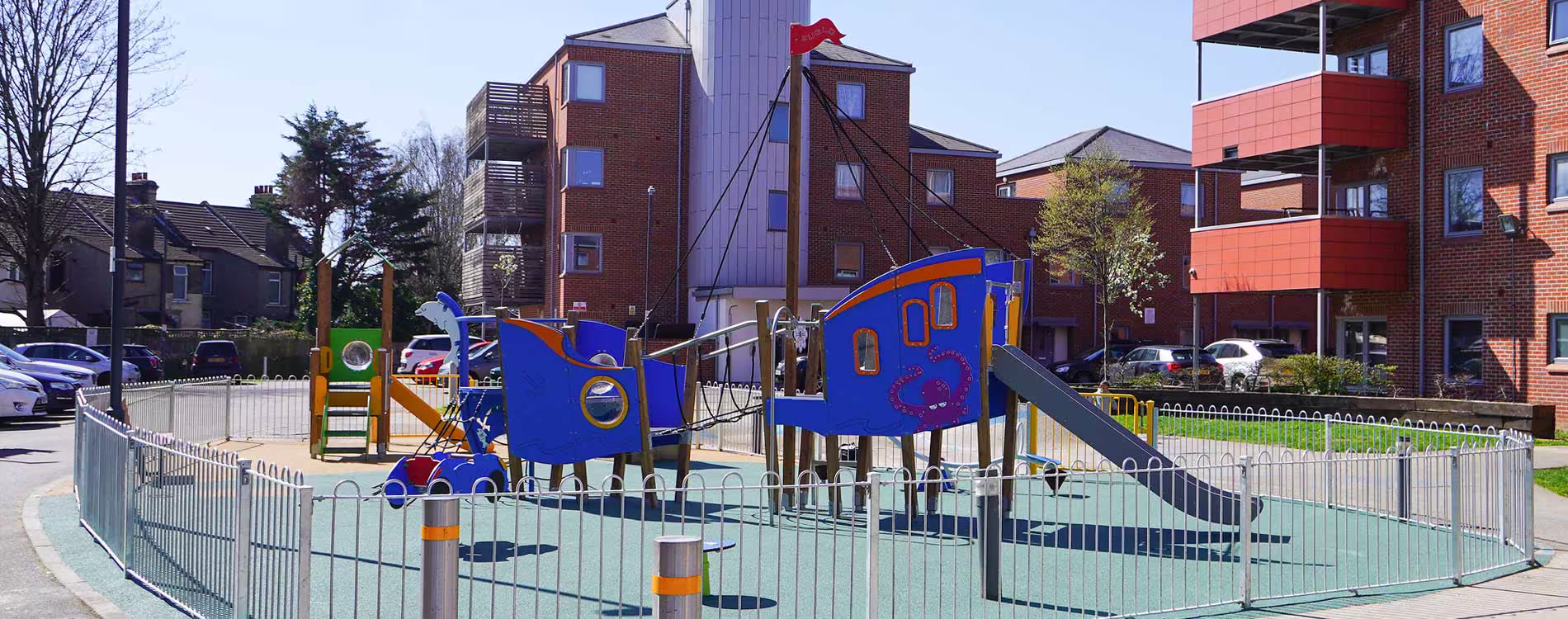 A creatively designed children's playground in a residential area, featuring a large, blue pirate ship climbing frame with a slide and climbing features.