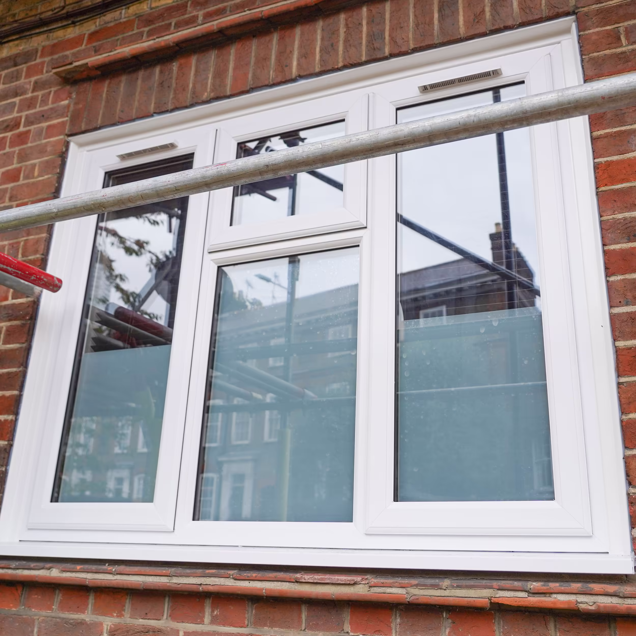 Close-up of a new white uPVC double-glazed window installed in a red brick wall, with a scaffolding pole in the foreground reflecting the ongoing works.