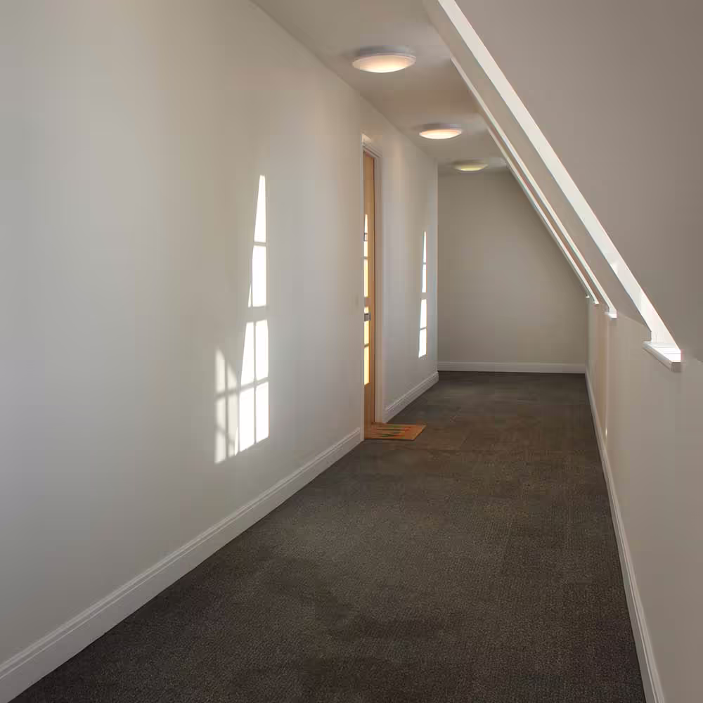 A newly refurbished communal corridor on the top floor of a building, featuring fresh white walls, new grey carpet, and modern lighting as part of a cyclical maintenance programme.