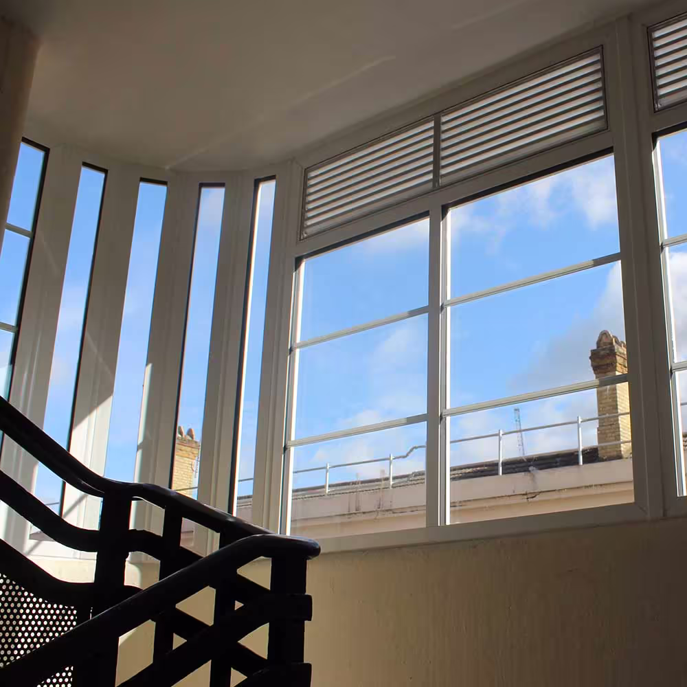 Interior of a redecorated stairwell in an Art Deco building, looking out through large restored windows with white frames onto a blue sky.