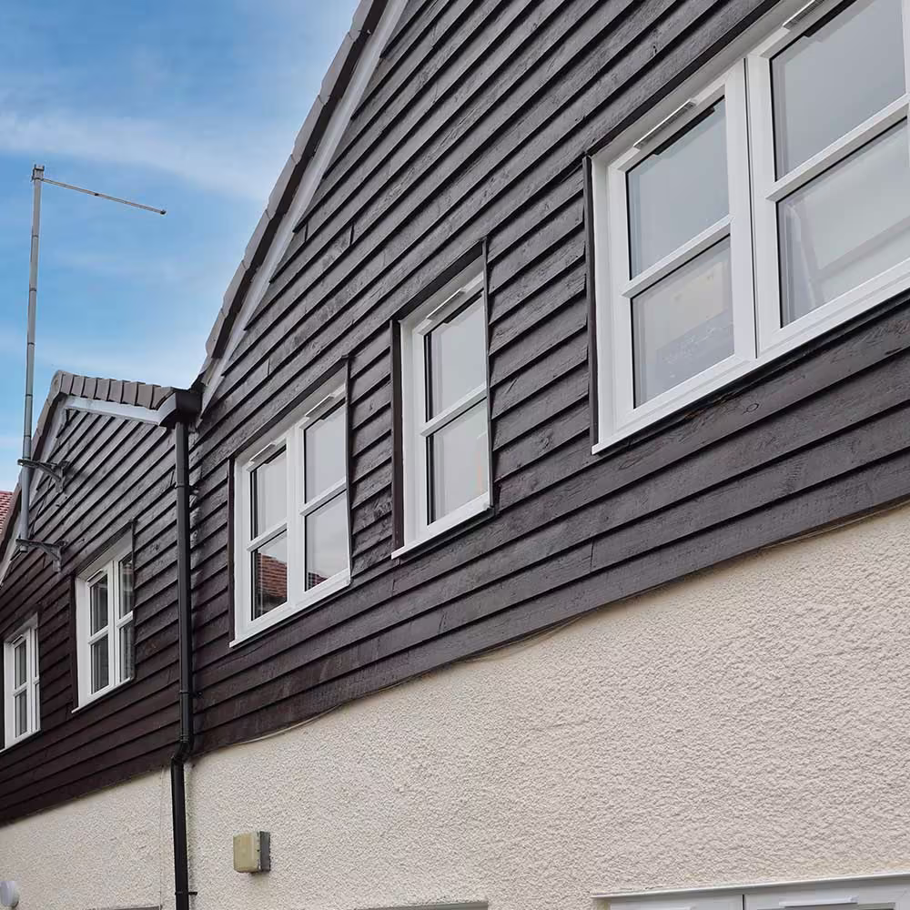 The facade of a property after external property services, featuring a contrast between the lower rendered wall and the dark brown weatherboarding above, with new white windows.