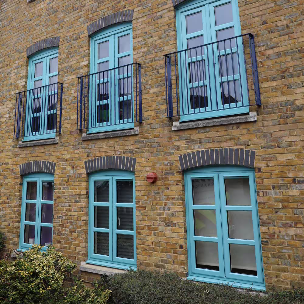 The yellow brick facade of a residential building featuring newly painted turquoise windows and dark blue Juliet balconies, showcasing comprehensive external property services.