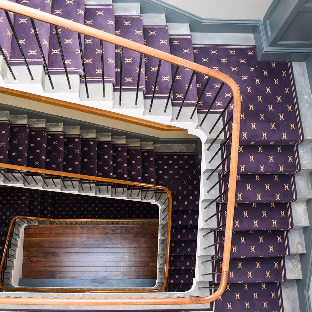 A top-down view of a grand, multi-flight staircase in a Grade Listed building, fitted with a new, elegant purple patterned carpet runner after repairs.