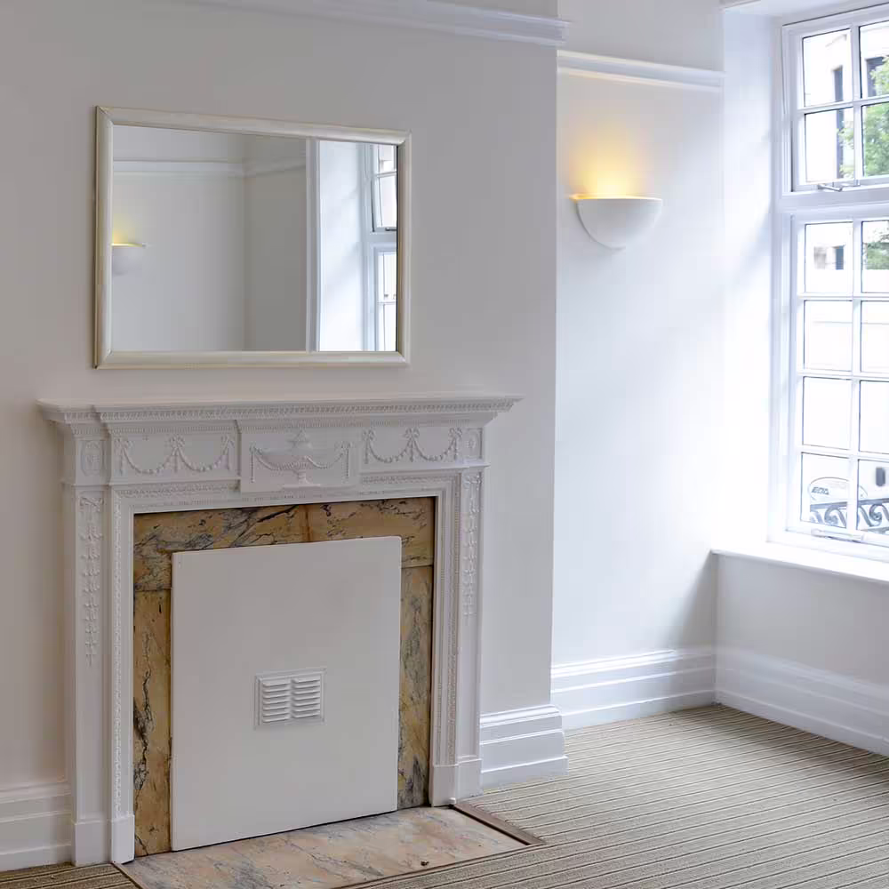 A fully refurbished room in a heritage building, featuring a conserved ornate white fireplace with a marble insert, set against clean white walls and new carpeting.