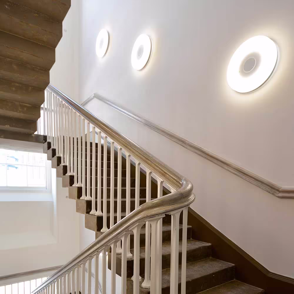 A grand traditional staircase in a renovated heritage building, blending original stone steps and a wooden handrail with modern circular wall lights.