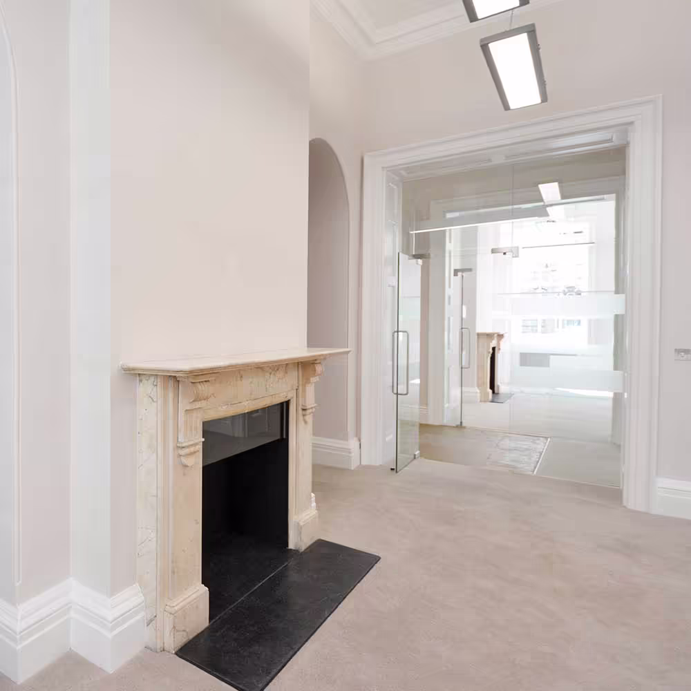 A sensitively renovated room in a heritage building, contrasting a classic marble fireplace with a modern, frameless glass door leading to an adjacent office space.