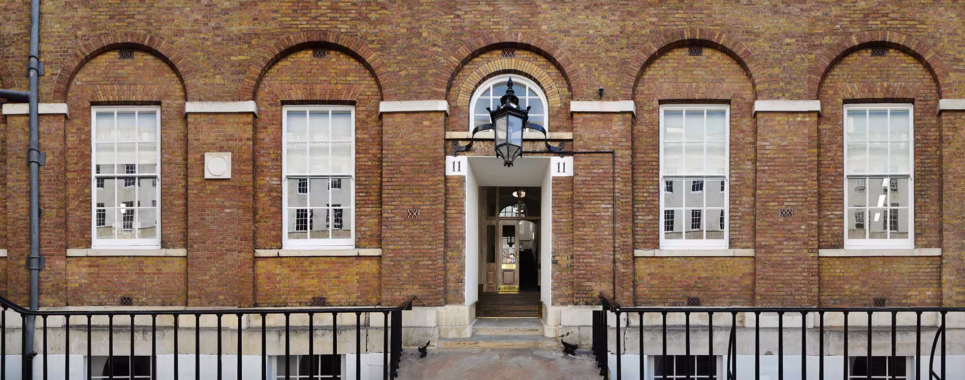 Symmetrical panoramic facade of a renovated heritage brick building, featuring large arched sash windows and a central arched entrance with a traditional lantern.