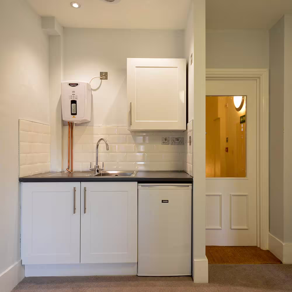 A compact and modern white kitchenette installed during a heritage void project, complete with a sink, fridge, and instant water heater.