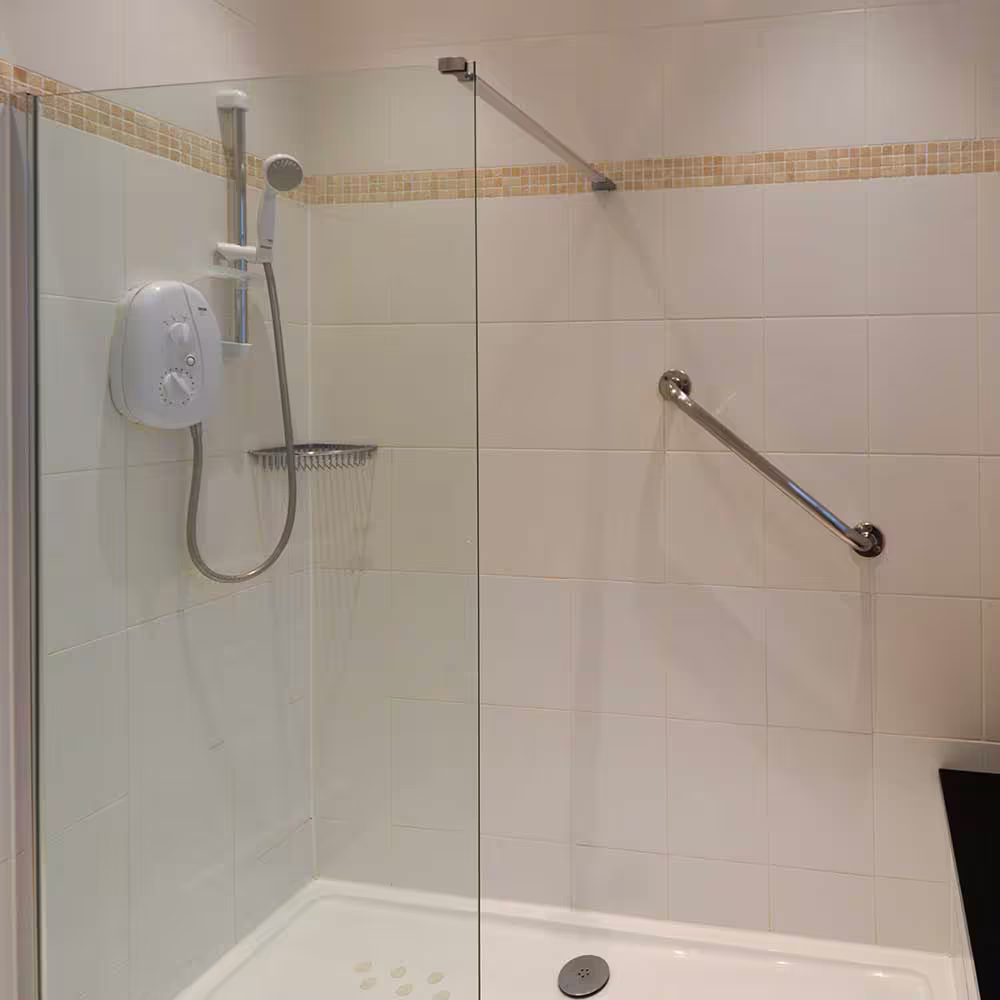A modern, accessible shower installed during a high-end refurbishment, featuring a walk-in shower tray, glass screen, white tiles, and a chrome safety grab rail.