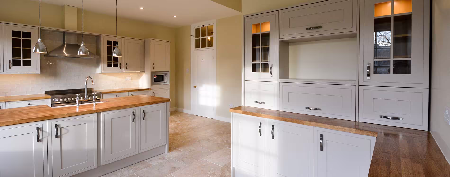 A panoramic view of a high-end kitchen refurbishment, featuring classic light grey shaker cabinets, warm oak worktops, and a central island with a sink.