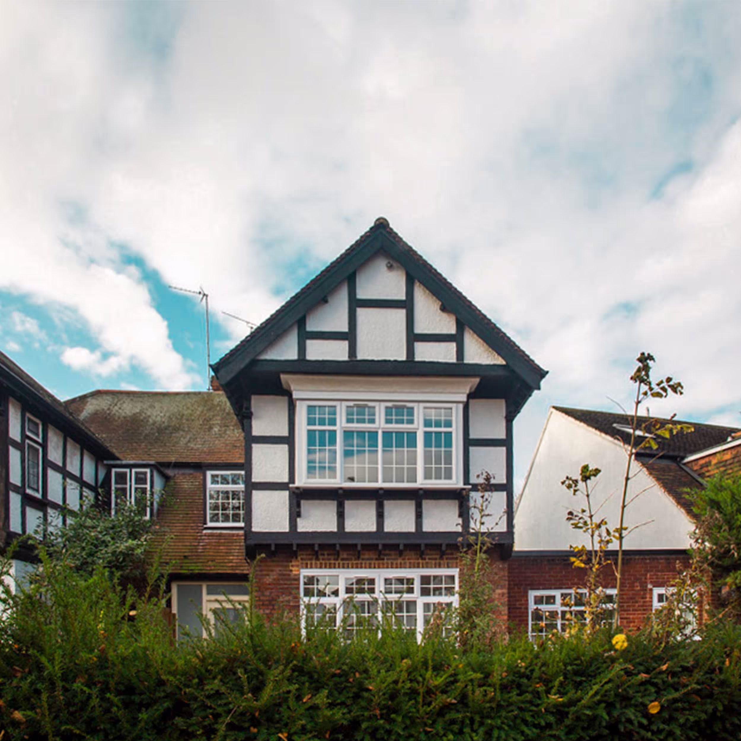 The exterior of a traditional half-timbered house which has undergone a housing conversion, viewed from the street over a tall green hedge.