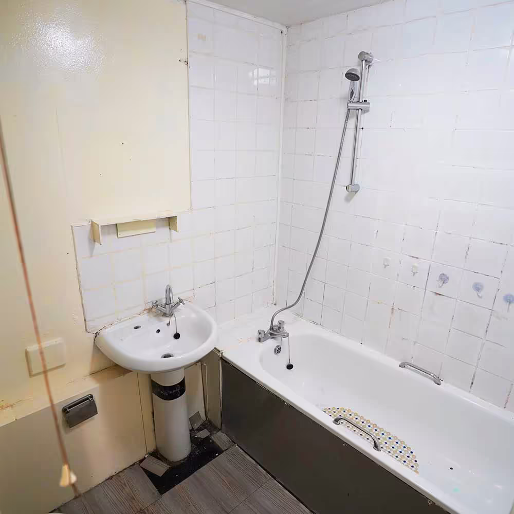 A worn-out bathroom in a council property before replacement, showing stained tiling, an old bath and a small pedestal sink.