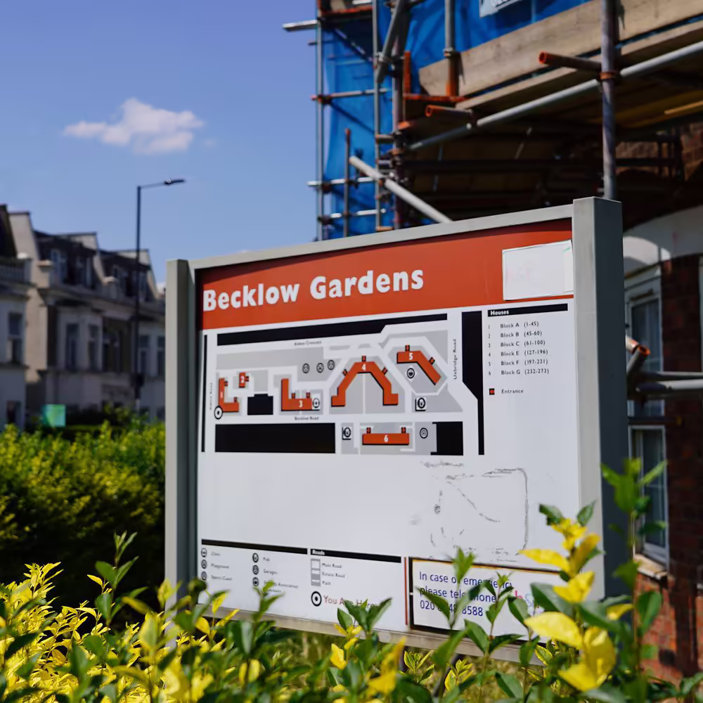 A site information board for the Becklow Gardens estate, with a building in the background covered in scaffolding as part of a major refurbishment project.