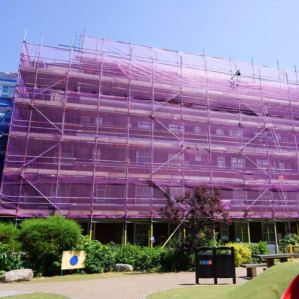 A large residential building completely shrouded in purple scaffolding and safety netting during a major refurbishment, viewed from a nearby park.