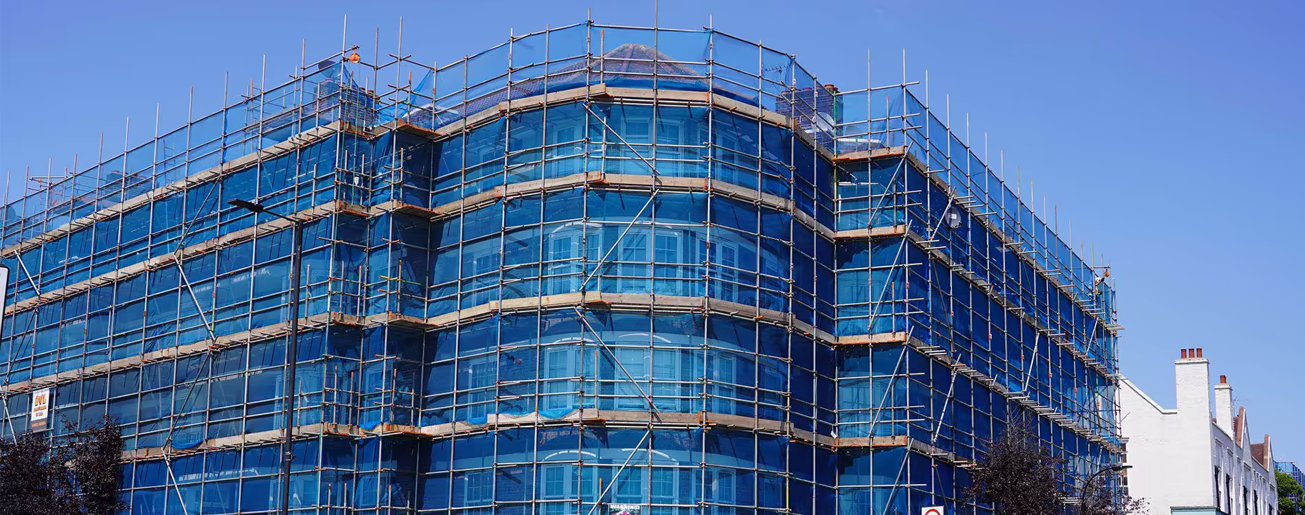 A large, multi-storey corner building undergoing major refurbishment works, completely enveloped in scaffolding and blue safety netting against a clear blue sky.