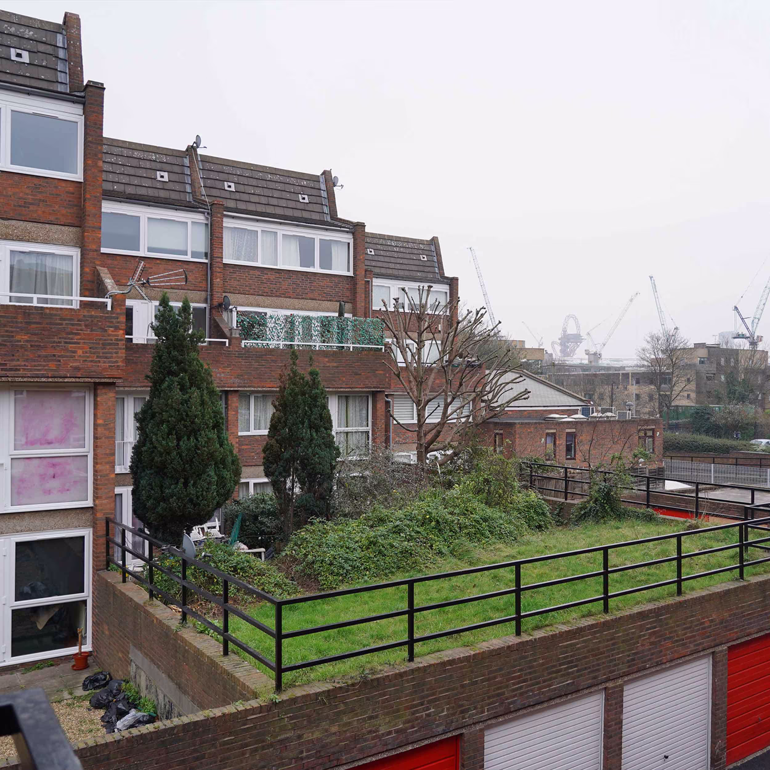 A view across the housing estate showing a communal rooftop garden with a lawn, and red garage doors below, part of the planned external works.