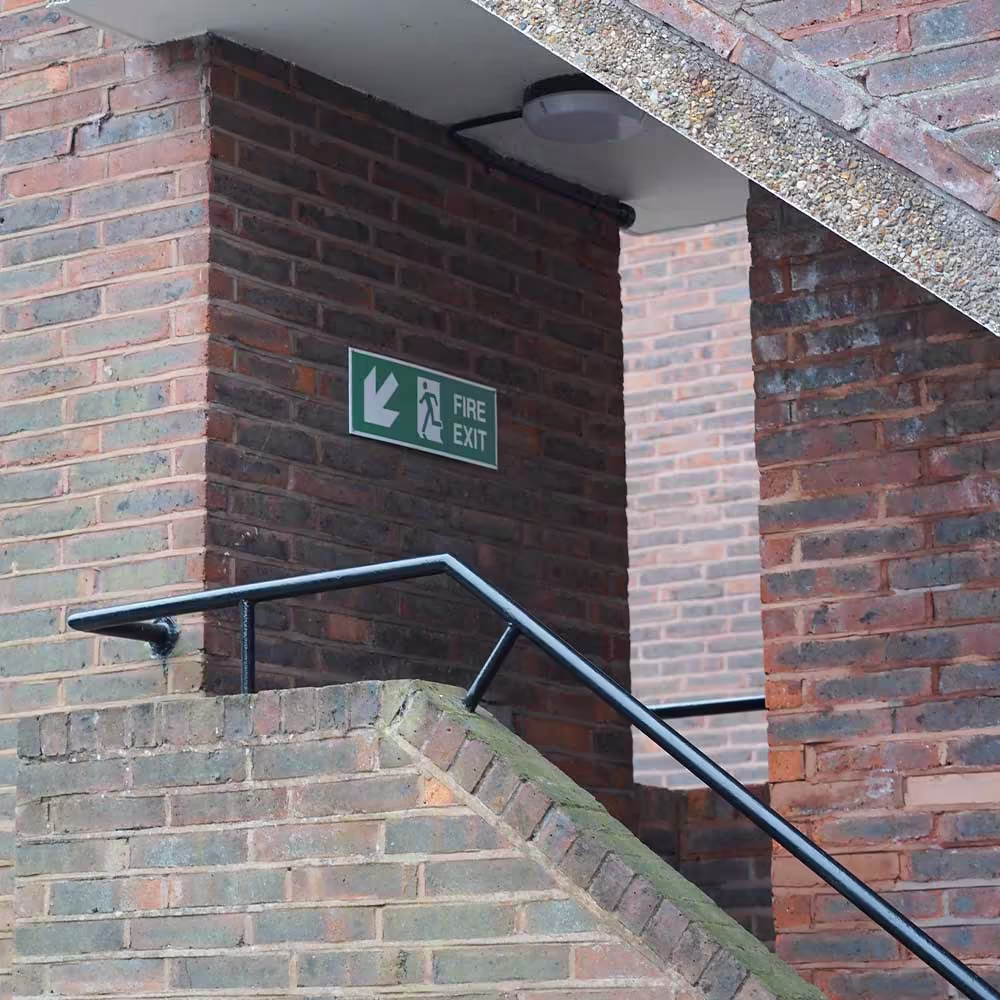 A newly installed black metal handrail in a brick communal stairwell, with a green fire exit sign, part of the planned safety and decoration works.