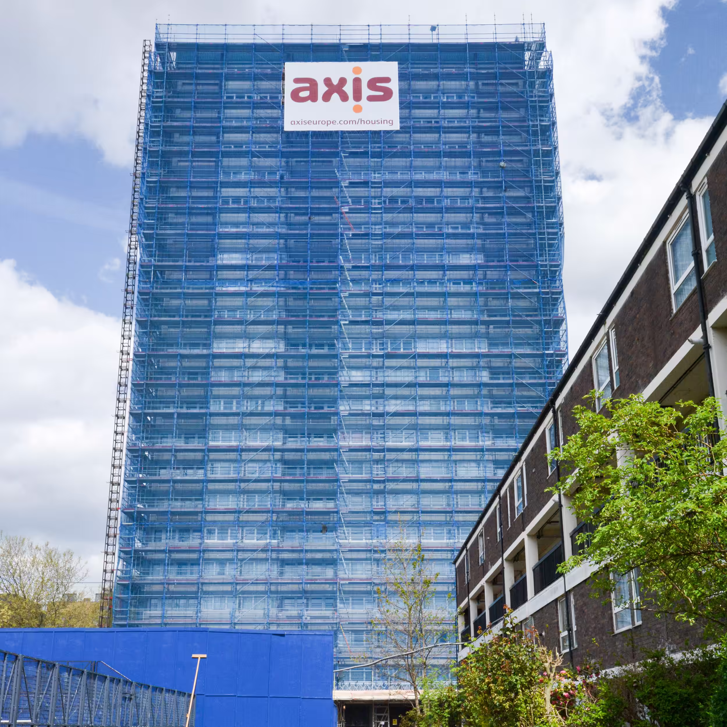 A tall residential tower block fully enveloped in blue scaffolding and netting, with a large Axis logo displayed at the top during a planned improvements project.