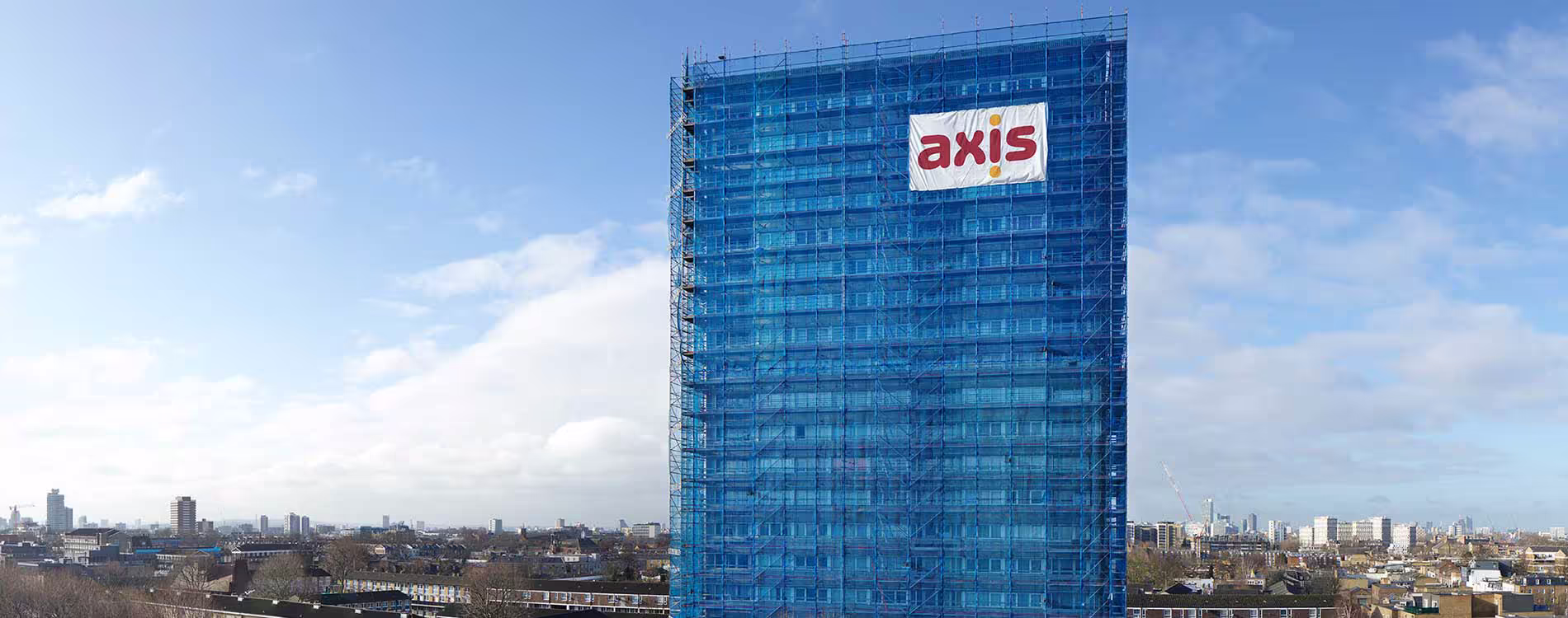 A panoramic view of a high-rise tower block completely wrapped in blue scaffolding with a large Axis logo, undergoing planned improvements against a city skyline.