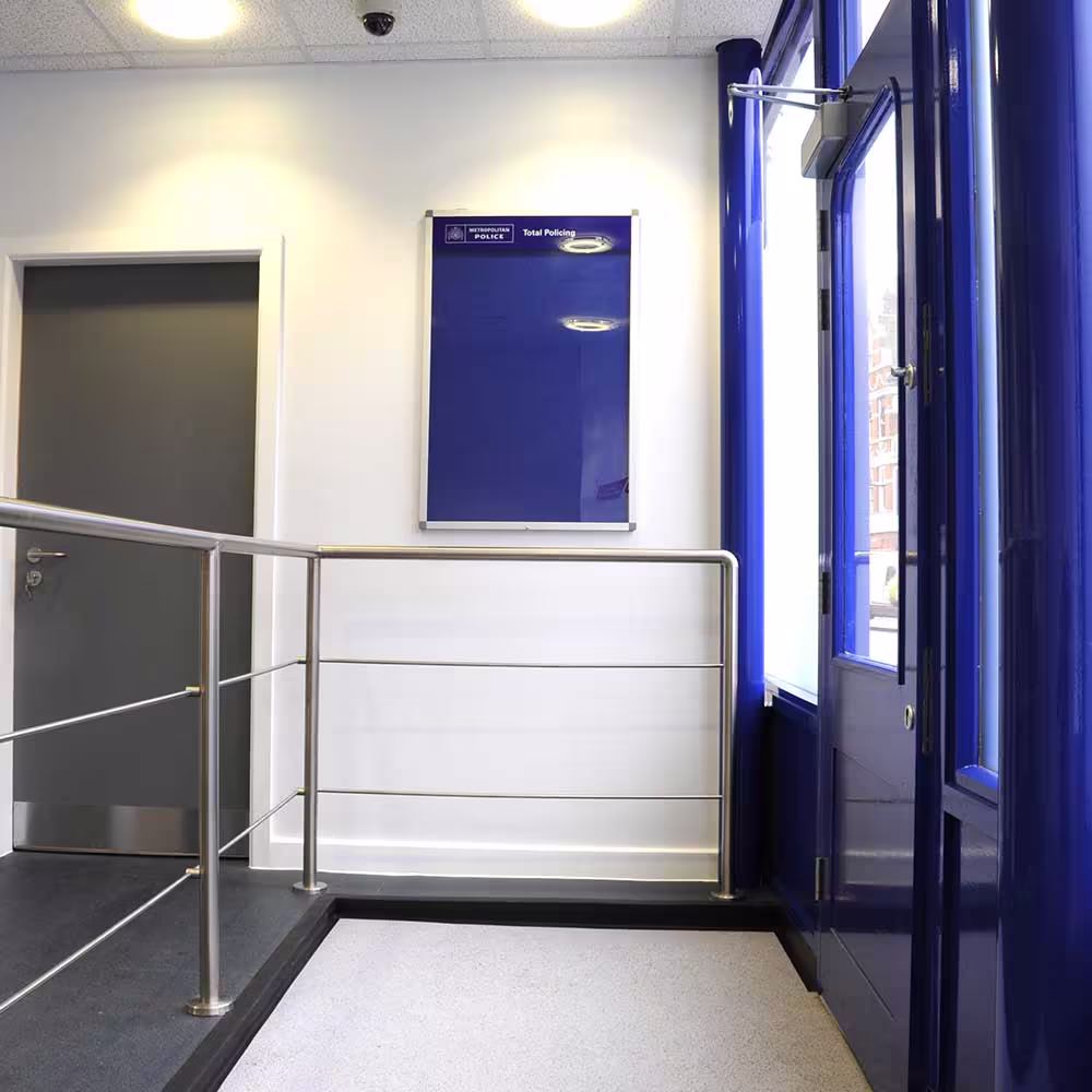 The modernised and accessible entrance area of a police building, featuring a bright blue door frame, a stainless steel handrail, and a Metropolitan Police noticeboard.