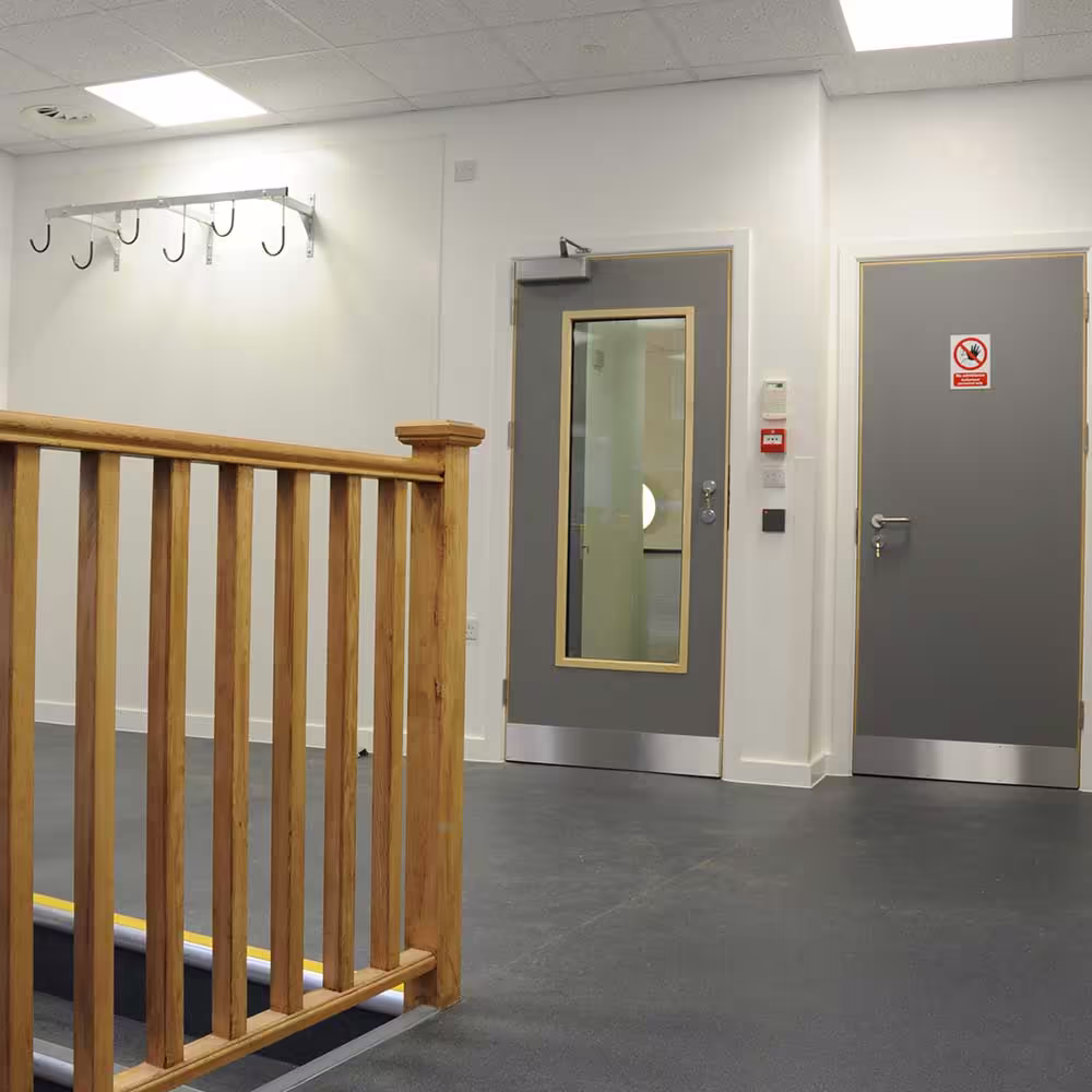 A refurbished corridor within the police building, showing new grey security doors with kick plates, durable dark flooring, and a wooden banister.
