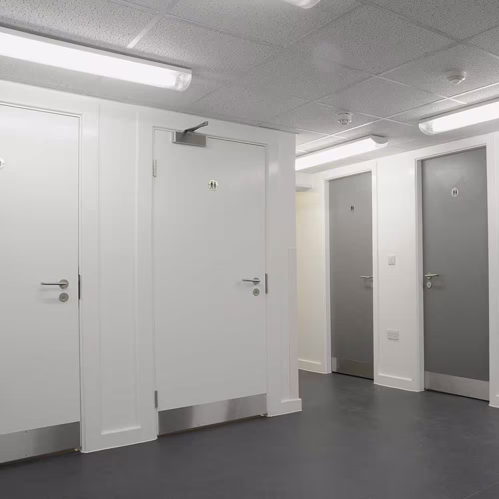 The fully refurbished communal toilet facilities in the police building, showing a row of modern, durable white and grey doors with stainless steel hardware.