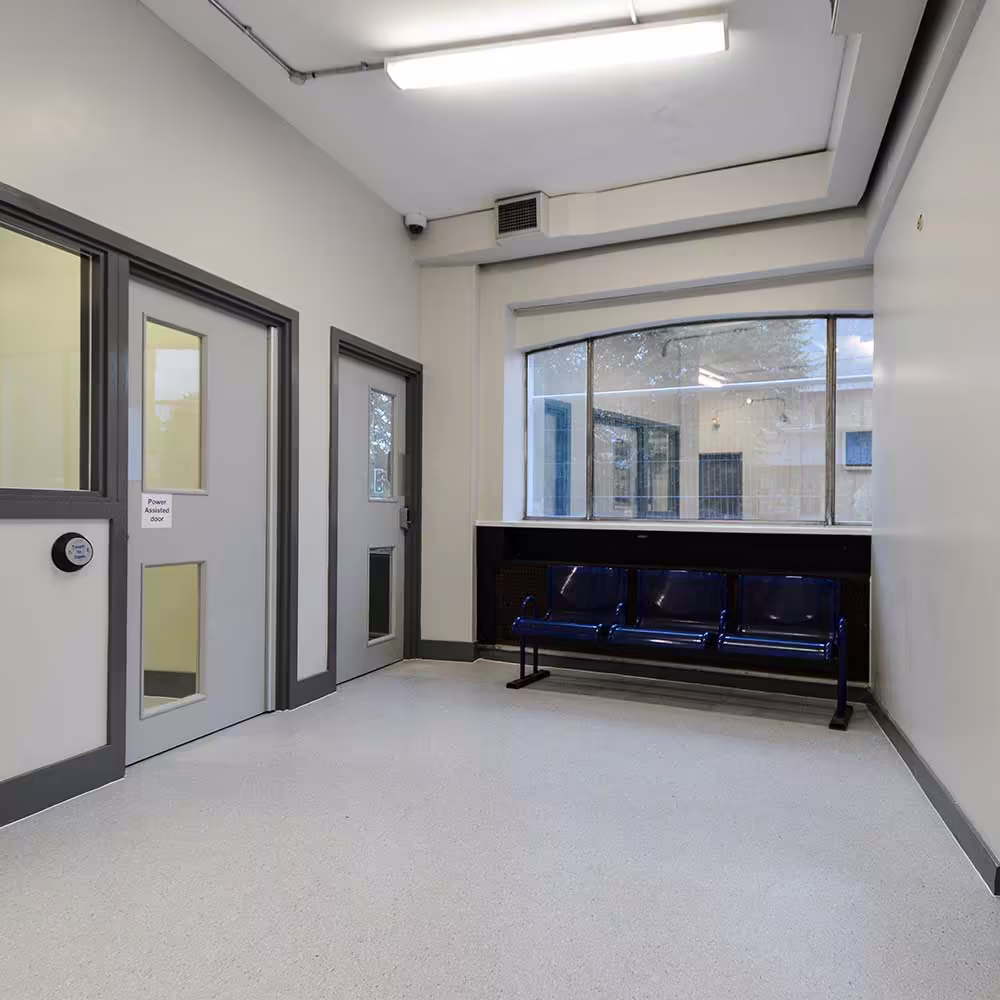 A wide view of the refurbished police station waiting area, showing the new secure doors, durable flooring, and blue public seating under a large window.